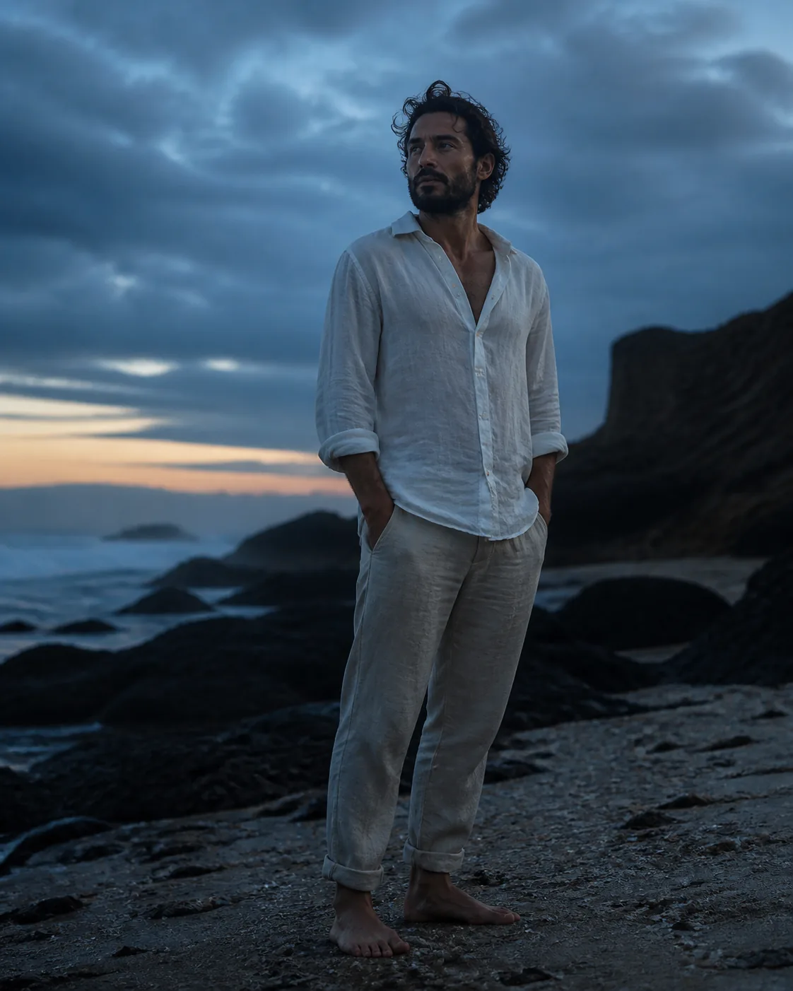 A fictional adult man in a white linen shirt standing on a quiet beach in a light sea breeze, soft golden-hour light, a natur