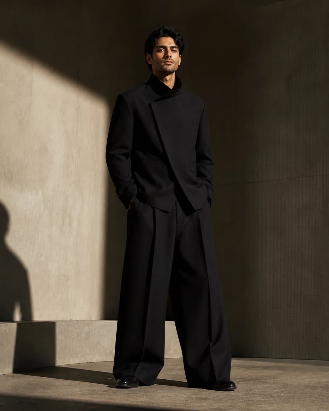 A fictional adult man with medium olive skin in his late 20s wearing a sculptural black jacket and wide-leg trousers in a min