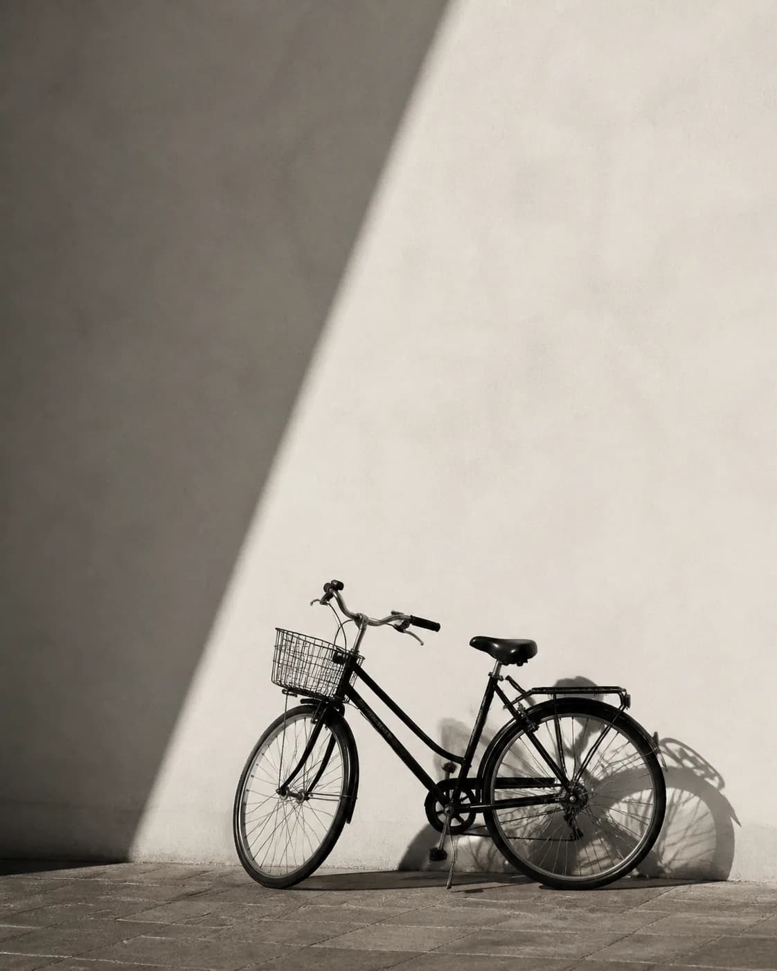 Minimalist monochrome photograph of a black city bicycle against a warm off-white wall with one crisp diagonal shadow, strong