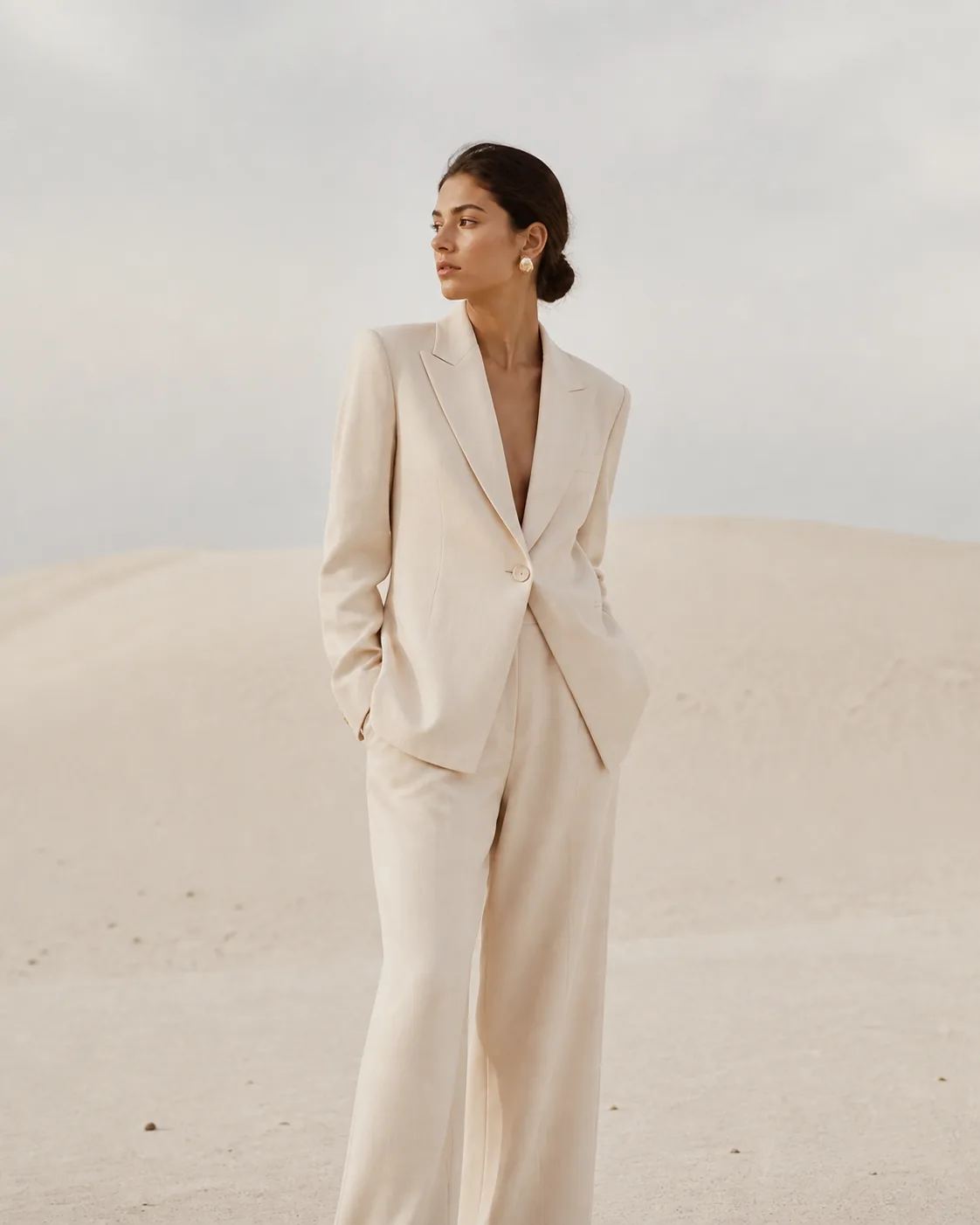 Minimalist editorial fashion photograph of a fictional adult woman in a structured ivory suit standing in a pale sand landsca