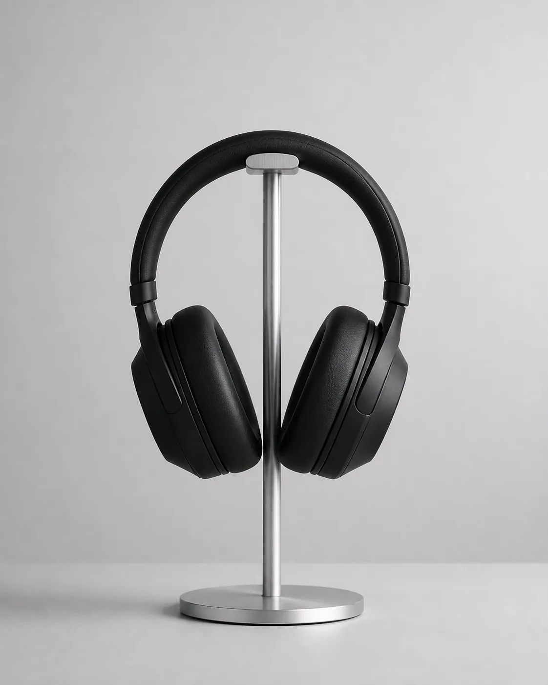 Minimalist product photograph of matte-black over-ear headphones on a thin brushed-metal stand, a pale-gray seamless backgrou